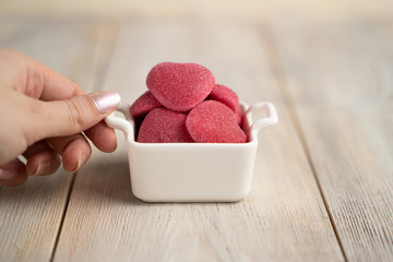 Marmalade pink hearts in a white plate-basket with a hand on a light beige wooden background, a concept for Valentine's Day, wedding, birthday and other holidays, a view from the top.