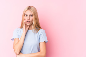 Young blonde woman on pink background is saying a secret hot braking news and looking aside