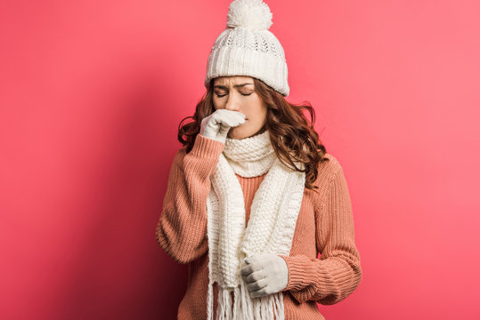 diseased girl in warm hat and scarf touching nose on pink background