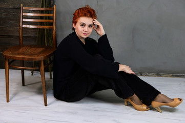 Fototapeta premium Art portrait of a pretty model with short red hair in dark clothes on a gray background in the interior. A woman sits next to a chair right in front of the camera in various poses.