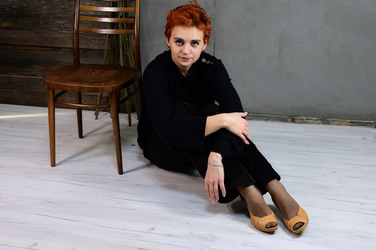 Art portrait of a pretty model with short red hair in dark clothes on a gray background in the interior. A woman sits next to a chair right in front of the camera in various poses.