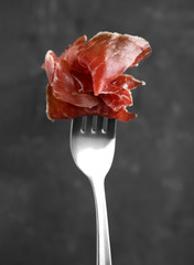Tasty prosciutto on fork against grey background