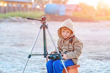 A boy with binoculars sits on a chair on the street, next to a tripod.