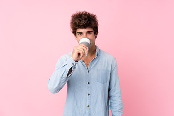 Young caucasian man over isolated pink background holding and drinking a coffee to take away