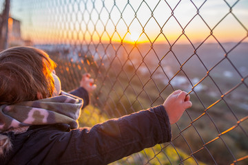 Kid boy holding hands on wire fence at sunset