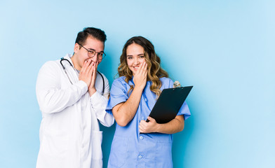 Young doctor couple posing in a blue background isolated laughing about something, covering mouth...