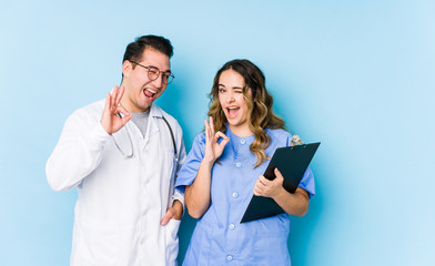 Young doctor couple posing in a blue background isolated winks an eye and holds an okay gesture...