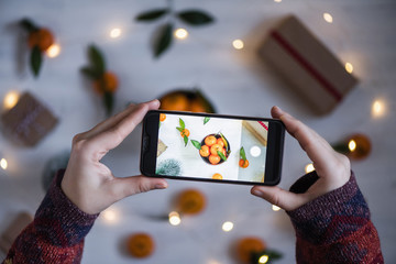 Woman blogger taking a picture on mobile phone her decoration of mandarin, Christmas tree, gifts, luminous garland on the white wooden boards background