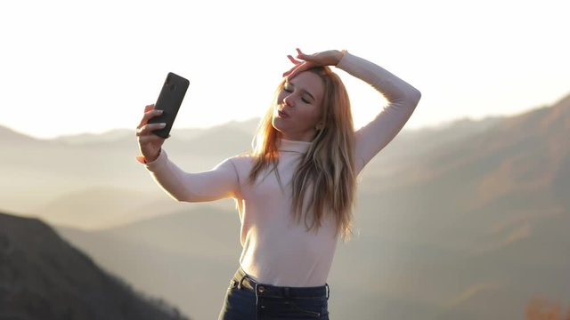 funny and happy young woman in white clothes stay on mountain top and do a selfie on phone . beautiful view on cloudy rock at sunset enjoying travel adventure