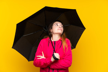 Young brunette girl holding an umbrella over isolated yellow background making doubts gesture while lifting the shoulders