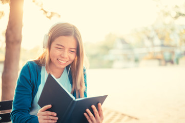 Smiling woman in eyeglasses reading book and listening to music with headphones in park outdoor against sunlight,City lifestyle concept.