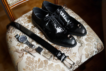 The groom is gathering in the morning. Gray chair. Men's Black classic patent leather shoes. Black belt. Wedding details. Eau de toilette, perfume, leather strap watch. Wedding rings.