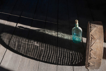 Rural still life with empty bottle and old sieve creating beautiful shadows on unpainted wooden surface. The bottle is closed with a paper cork.