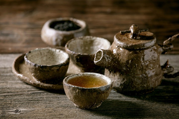 Hot black tea in traditional fireclay ceramic craft cups served with plate, teapot and bowl of dry tea leaves on old wooden table. Dark rustic atmosphere.