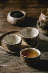 Hot black tea in traditional fireclay ceramic craft cups served with plate, teapot and bowl of dry tea leaves on old wooden table. Dark rustic atmosphere.