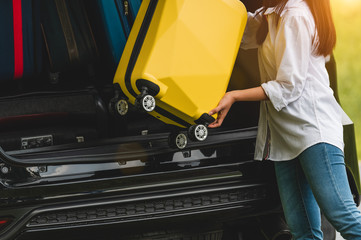 Asian woman lifting yellow suitcase into SUV car during travel in long weekend trip. People lifestyles and transportation concept. Girl put luggage from car trunk to vacation camping in summer time - obrazy, fototapety, plakaty