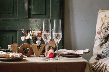 st. Valentines day or wedding romantic table setting with wooden letters love, needle felted hearts, ceramic plates, bouquet of pink roses, couple glasses of champagne on natural linen tablecloth.