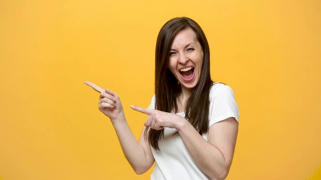 Beautiful cute brunette young woman in white t-shirt showing thumbs up, pointing on copy space with charming smile isolated over yellow background in studio. People emotions lifestyle concept. Mock up