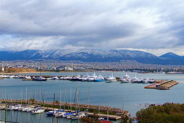 Fototapeta na wymiar Aerial winter landscape view of harbor with moored luxury yacht and boats in marina of the city of Piraeus. Mountain rang in snow in the background. Attica, Greece