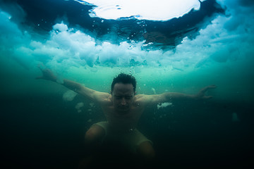 Young man swims in the ice hole made on the winter lake. Underwater view with the ice floating in...