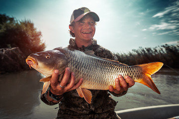 Happy young fisherman holds the big Carp fish (Cyprinus carpio) and smiles - obrazy, fototapety, plakaty