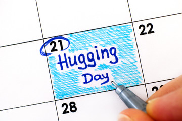 Woman fingers with pen writing reminder Hugging Day in calendar.