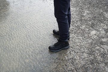 A boy in waterproof shoes standing in the border between a puddle and gravel ground. the tips of the shoes pointing to the puddle water 