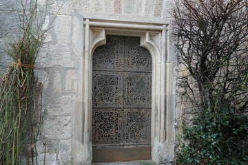 Fototapeta na wymiar Metal entrance door with smithing or wrought metal decoration in a white stone wall surrounded by leafless bushes, ivy and rose bushes in winter, like in a castle of Sleeping Beauty. 