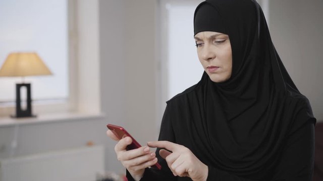 Side view portrait of young Muslim woman scrolling on smartphone and gesturing no by shaking head. Modern eastern lady shocked with social media. Traditional culture, society.