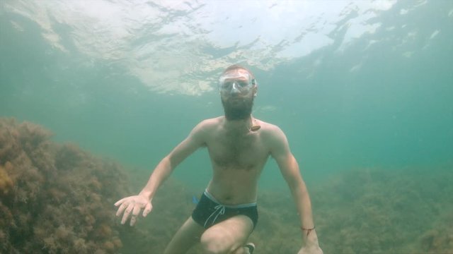 A thin man with a beard in a mask swims underwater, waving his arms. Close-up.