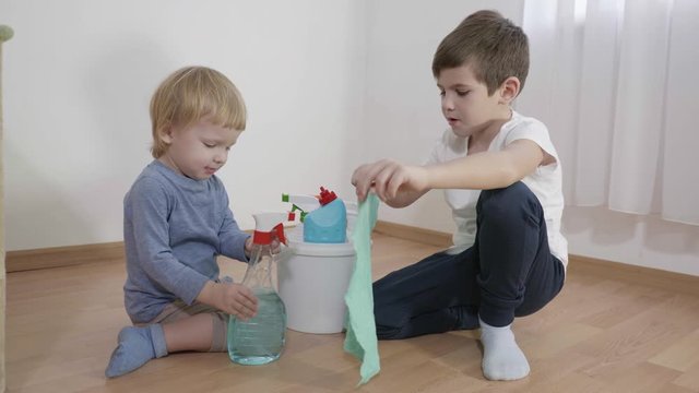 relationship brothers, little boy helps his older brother during cleaning, a child with a plastic bottle sprays detergent on a rag sitting on the floor at home
