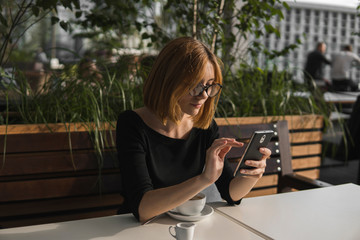 Young business woman uses phone. Good looking brunette female uses online banking on smart phone to transfer money from credit card. Girl using smart phone and chatting with business partners in cafe.