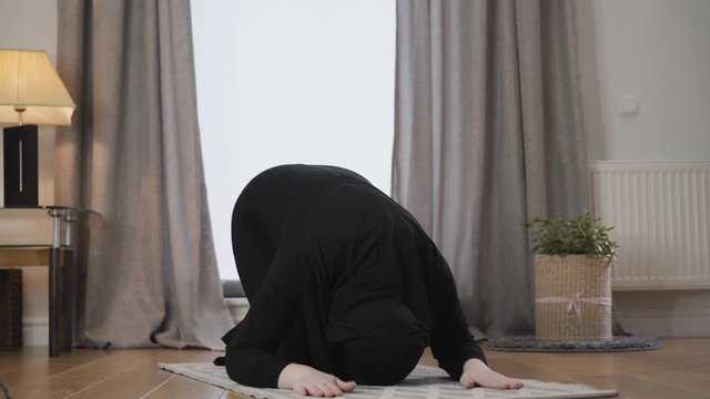 Young Muslim lady in black traditional hijab bowing down and raising up with hands raised. Modern eastern woman praying at home. Eastern culture, religion, lifestyle.