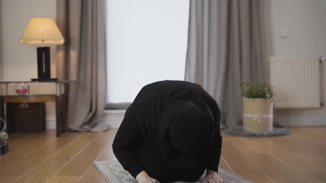 Portrait of young Muslim woman in black traditional hijab bowing down and raising up. Eastern woman praying at home. Eastern culture, religion, lifestyle.