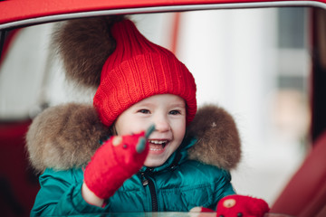 Smiling cute winter girl in red hat sitting in car having fun medium shot. Happy beautiful female baby in warm clothing having positive emotion outdoor surrounded by snowflakes enjoying childhood