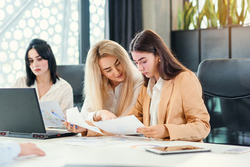 Likable young female office workers sitting at the meeting room and discussing their joint business project using financial datas and computer.