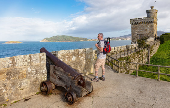 A hiker looks at the Atlantic from a castle in Galicia in northern Spain. There is a historic cannon in the foreground and a watchtower in the background. Sunshine and blue sky.