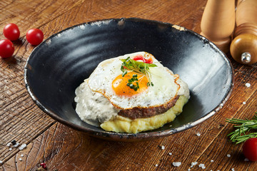 Close up view on Beef steak with fried eggs, mushroom cream sauce and mashed potatoes in a black bowl in a composition with spices on a wooden background. Tasty food