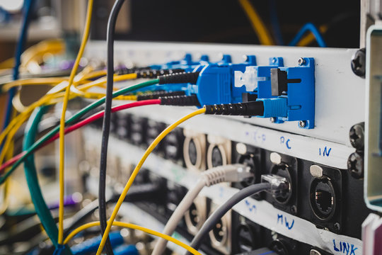 Different BNC SDI UTP and optical signal cables plugged in the back of a patch panel. A mess of cables visible behind the rack or panel. Focus on the  blue optical connectors.