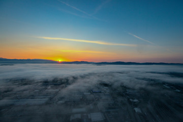 Aerial view of a foggy sunrise over a residential and industrial area of a city. Fog layered over houses below early morning sunrise.