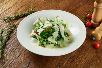 Tasty salad with lettuce, arugula, parmesan and cherry tomatoes in a white bowl on a wooden background in a composition with spices