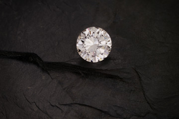 A solitaire, faceted, clear, round diamond sits on a dark rough slate background.
