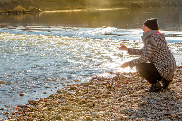 Lady crouches at the shore of the river in a winter cold morning and playfully throwing pebbles in the water.