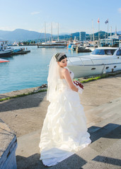 Fototapeta na wymiar Wedding by the sea. Bride with a red bouquet near sea port. Mountains and sea background. Bride with crown and veil. Just married in Batumi, Georgia.