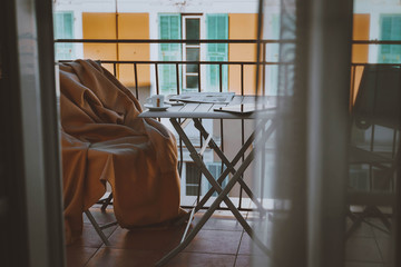 cozy terrace or balcony with small table, chair with blanket, newspaper, phone, tablet, small cup of coffee on the table. Yellow buildigs with big french windows on background.