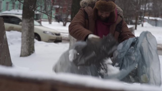 a bearded homeless man in a hat earflaps and fur coat rummages in the trash