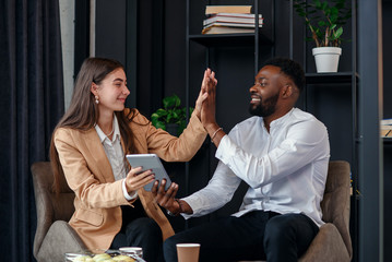 African man and caucasian woman have coffee break at office rest room at table near window drinking hot espresso.
