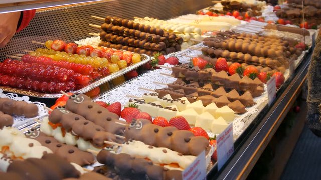 Various fruits on a wooden skewer stick in a caramelized glaze made of milk black white chocolate. Grapes, banana, strawberries in icing on the counter of the Christmas market