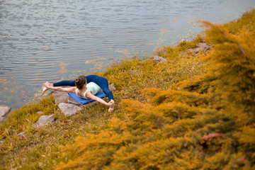 Fototapeta na wymiar A young sports girl practices yoga on a fall yellow lawn by the river, use yoga assans posture. Meditation and unity with nature