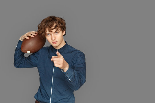 Sporty Lifestyle. Teenager boy standing isolated on grey with american football ball pointing at camera cool space for text or product - Powered by Adobe